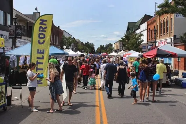 A bustling street fair in downtown Alliston with people walking among vendor booths. A bright yellow "Kettle Corn" sign stands out. The atmosphere is lively and vibrant.