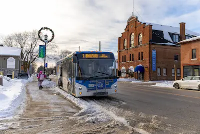 A city bus stopped on a snowy street with a brick pharmacy building nearby. A person in a pink jacket walks along the icy sidewalk, under a festive wreath.