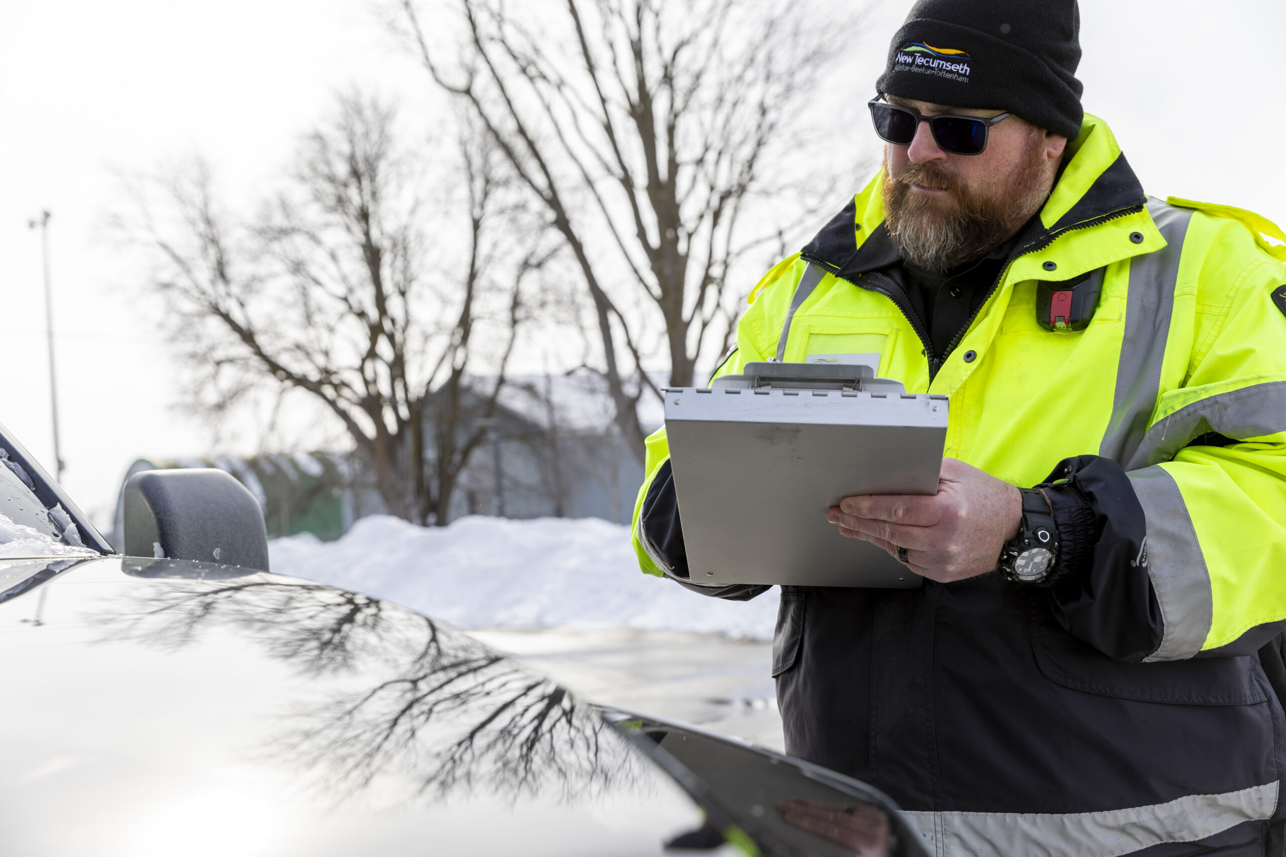 A man in a bright reflective jacket places a parking ticket on a vehicle's snow-covered windshield. He appears focused. Bare trees in the background.