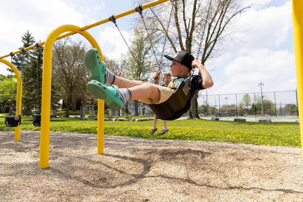 A child swinging on a playground swing with another child playing in the background.