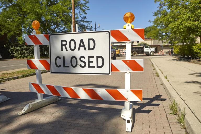 Road closure barricade with orange warning lights and a “Road Closed” sign blocking a street, with construction vehicles and work zone equipment visible in the background on a sunny day.
