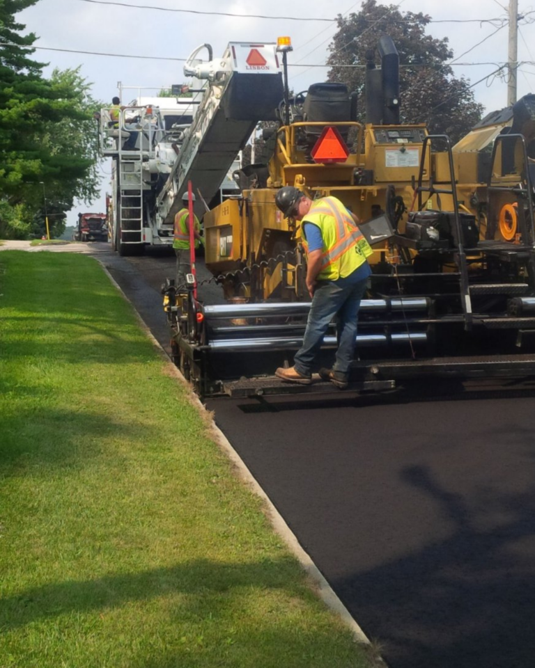 Road paving crew resurfacing a residential street with paving equipment. Fresh black asphalt is being laid along the roadway while workers in high-visibility safety vests operate machinery and monitor the paving process. Grass boulevards, mature trees, utility lines, and residential properties are visible along the street.