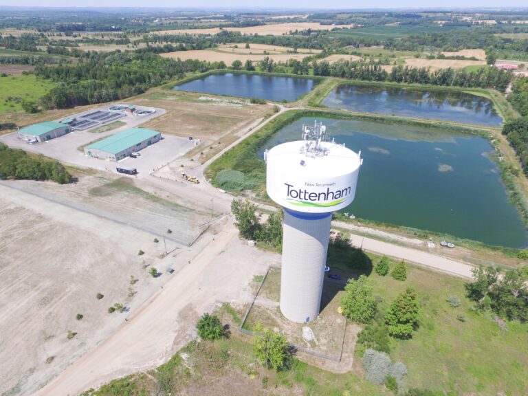 Aerial view of the Tottenham Wastewater Treatment Plant in New Tecumseth, featuring a large water tower marked "Tottenham," treatment buildings, and several wastewater lagoons surrounded by agricultural fields, trees, and rural landscape.